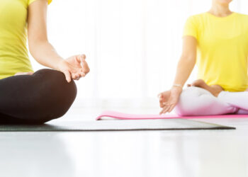 Duas pessoas vestindo camisas amarelas sentam-se de pernas cruzadas em tapetes de ioga em um ambiente interno, praticando meditação com as mãos apoiadas nos joelhos, tocando as pontas dos dedos. A cena é clara com um fundo branco e suave. | YogaLounge