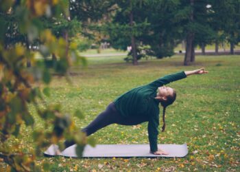 Uma pessoa pratica yoga e saúde cerebral ao ar livre em um tapete, fazendo uma pose de ângulo lateral com um braço estendido acima da cabeça, cercada por grama, árvores e folhas de outono espalhadas. | YogaLounge