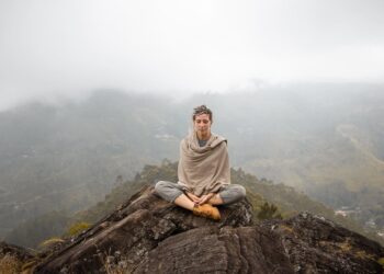 Uma pessoa senta-se de pernas cruzadas no topo de uma colina rochosa, meditando com os olhos fechados com bem-estar consciente. Ela usa roupas de cor neutra e um xale. Montanhas enevoadas e um vale com casas espalhadas podem ser vistos ao fundo. | YogaLounge