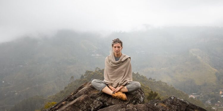 Uma pessoa senta-se de pernas cruzadas no topo de uma colina rochosa, meditando com os olhos fechados com bem-estar consciente. Ela usa roupas de cor neutra e um xale. Montanhas enevoadas e um vale com casas espalhadas podem ser vistos ao fundo. | YogaLounge