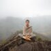 Uma pessoa senta-se de pernas cruzadas no topo de uma colina rochosa, meditando com os olhos fechados com bem-estar consciente. Ela usa roupas de cor neutra e um xale. Montanhas enevoadas e um vale com casas espalhadas podem ser vistos ao fundo. | YogaLounge