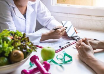 Um profissional da área de saúde discute planos de saúde com uma pessoa em uma mesa, cercada por uma prancheta, um smartphone, legumes, uma maçã verde, halteres cor-de-rosa e uma fita métrica - inspiração perfeita para quem participa de um seminário de coaching. | YogaLounge