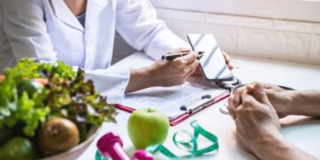 Um profissional da área de saúde discute planos de saúde com uma pessoa em uma mesa, cercada por uma prancheta, um smartphone, legumes, uma maçã verde, halteres cor-de-rosa e uma fita métrica - inspiração perfeita para quem participa de um seminário de coaching. | YogaLounge