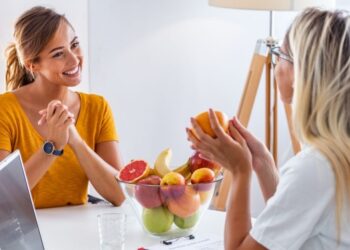 Duas mulheres sentam-se uma em frente à outra em uma mesa, conversando amigavelmente. Uma tigela de frutas e um laptop sugerem que elas estão discutindo insights de um seminário de coaching, enquanto uma mulher segura uma laranja e a outra sorri calorosamente. | YogaLounge