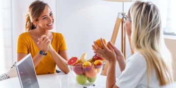 Duas mulheres sentam-se uma em frente à outra em uma mesa, conversando amigavelmente. Uma tigela de frutas e um laptop sugerem que elas estão discutindo insights de um seminário de coaching, enquanto uma mulher segura uma laranja e a outra sorri calorosamente. | YogaLounge