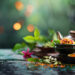 Uma configuração serena com um bule de chá, uma xícara, um incenso aceso, ervas frescas, especiarias e uma tigela com uma flor vibrante, todos dispostos em uma superfície refletora com um fundo verde exuberante e luzes bokeh quentes. | YogaLounge