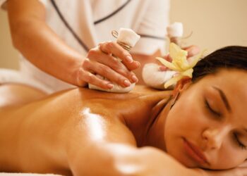 Uma mulher está deitada de bruços com os olhos fechados, recebendo uma massagem relaxante com compressas de ervas em suas costas. Ela tem uma flor amarela enfiada atrás da orelha e o ambiente parece calmo e de spa. Massagem ayurveda para saúde mental | YogaLounge