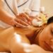 Uma mulher está deitada de bruços com os olhos fechados, recebendo uma massagem relaxante com compressas de ervas em suas costas. Ela tem uma flor amarela enfiada atrás da orelha e o ambiente parece calmo e de spa. Massagem ayurveda para saúde mental | YogaLounge