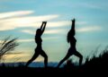 Duas pessoas estão em silhueta contra um céu azul e amarelo, praticando ioga ou poses de alongamento ao ar livre entre a grama alta. | YogaLounge