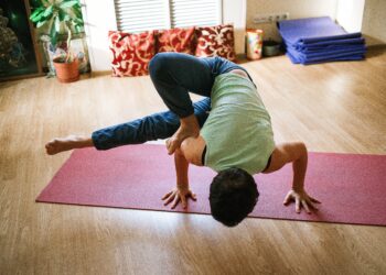 Uma pessoa pratica uma pose de ioga desafiadora em um tapete vermelho dentro de casa, equilibrando-se sobre as mãos com as pernas dobradas e um pé enganchado sobre o braço oposto. Adereços de ioga e plantas são visíveis ao fundo. | YogaLounge
