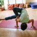 Uma pessoa pratica uma pose de ioga desafiadora em um tapete vermelho dentro de casa, equilibrando-se sobre as mãos com as pernas dobradas e um pé enganchado sobre o braço oposto. Adereços de ioga e plantas são visíveis ao fundo. | YogaLounge