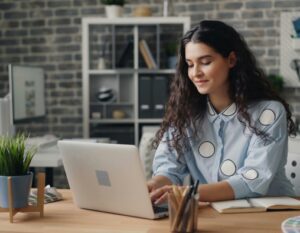 Uma mulher com longos cabelos cacheados, vestindo uma camisa azul clara com grandes pontos brancos, senta-se em uma mesa digitando em um laptop em um escritório moderno com prateleiras e plantas, pesquisando sobre a academia de yoga de Portugal. | YogaLounge
