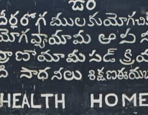 Uma placa preta com escrita Telugu branca e texto em inglês parcialmente visível na parte inferior, onde se lê "HEALTH HOME", fica do lado de fora de uma academia de ioga em Portugal, convidando os transeuntes a explorar o bem-estar. | YogaLounge