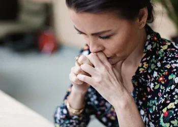Uma mulher vestindo uma blusa floral está sentada com as mãos fechadas perto da boca, olhando para baixo pensativamente, incorporando a intensidade silenciosa da ansiedade nas mulheres. | YogaLounge