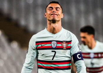 O jogador de futebol Cristiano Ronaldo, vestindo a camisa da seleção de Portugal, branca e listrada, com o número 7 e a braçadeira de capitão, está de pé com os olhos fechados e os lábios apertados — parecendo frustrado durante a partida, como se buscasse calma por meio da ioga. Outro jogador aparece ao fundo. | YogaLounge