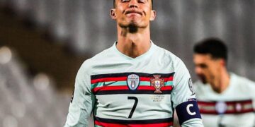 O jogador de futebol Cristiano Ronaldo, vestindo a camisa da seleção de Portugal, branca e listrada, com o número 7 e a braçadeira de capitão, está de pé com os olhos fechados e os lábios apertados — parecendo frustrado durante a partida, como se buscasse calma por meio da ioga. Outro jogador aparece ao fundo. | YogaLounge