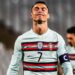 O jogador de futebol Cristiano Ronaldo, vestindo a camisa da seleção de Portugal, branca e listrada, com o número 7 e a braçadeira de capitão, está de pé com os olhos fechados e os lábios apertados — parecendo frustrado durante a partida, como se buscasse calma por meio da ioga. Outro jogador aparece ao fundo. | YogaLounge