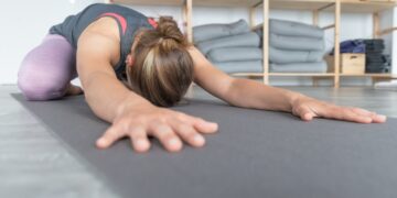 Uma mulher pratica ioga em casa, alongando-se para a frente sobre um tapete na postura da criança. Ao fundo, podem-se ver prateleiras com cobertores e almofadas dobrados, criando um espaço tranquilo para celebrar o Dia Internacional do Ioga. | YogaLounge